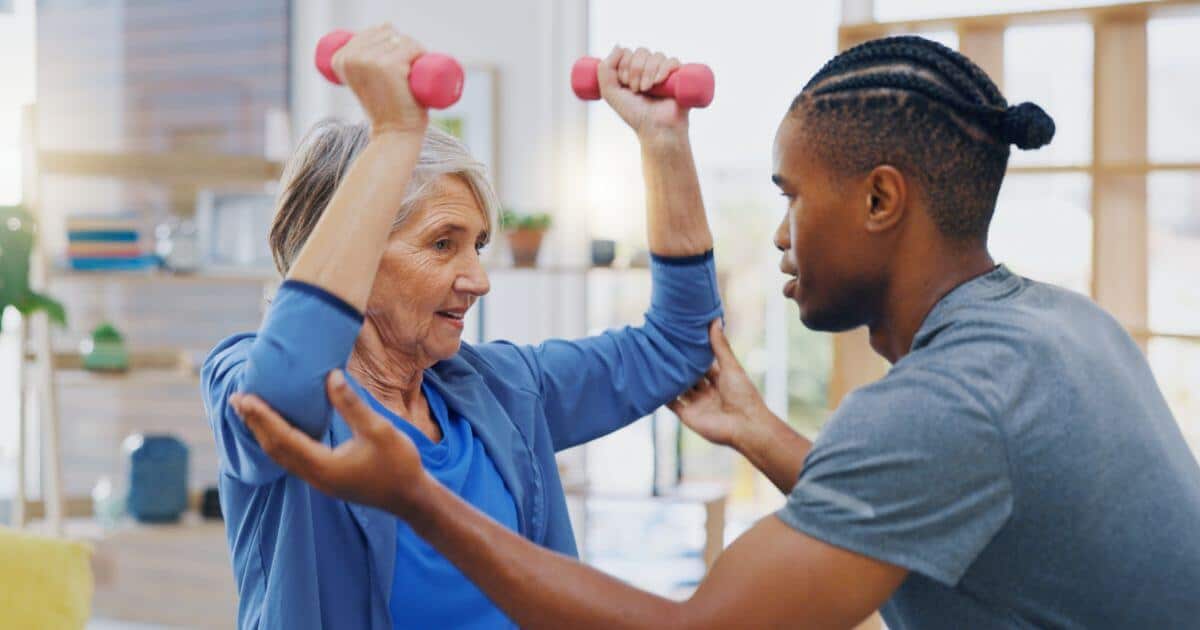 Elder woman in a fitness class for seniors lifting waits with a caregivers assistance.
