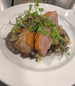 Image of white plate with sliced duck breast on top of mushroom sauce, topped with microgreens