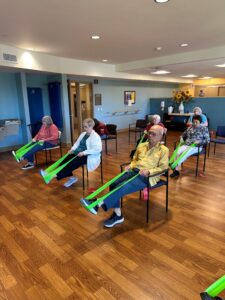 Older adults sit in chairs in fitness studio with green resistance bands around one foot, stretching the back of their legs