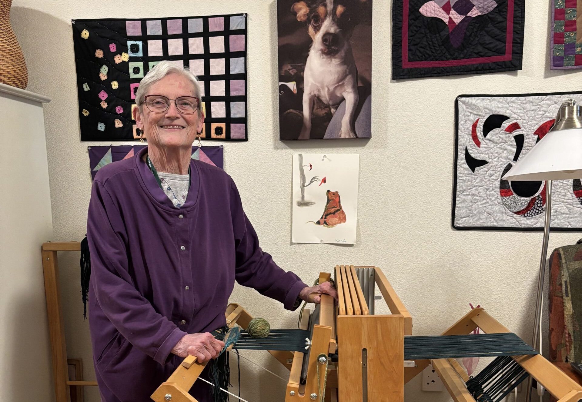 A woman stands beside a wooden loom with quilted artwork displayed on the wall behind her.